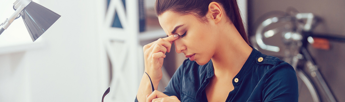 Zmęczone oczy – kobieta przy komputerze z zamkniętymi oczami trzyma się za nasadę nosa